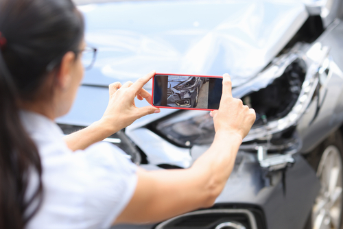 A woman uses her smartphone to take a photo of a car with significant front-end damage, documenting the aftermath of an accident.