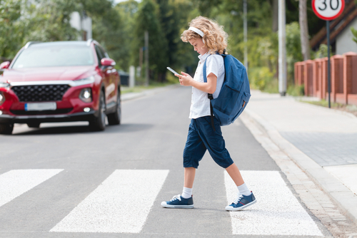 A young child with curly hair, wearing a blue backpack and casual clothes, is crossing a street while looking down at a phone. The child is walking on a marked pedestrian crosswalk.