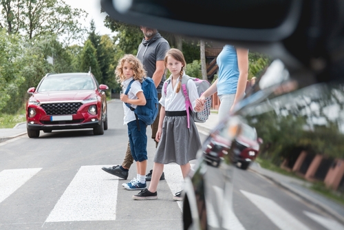 Two young children with backpacks and their guardians wait at a pedestrian crosswalk as a car approaches, highlighting the importance of road safety.