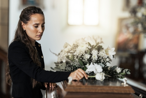 A grieving woman placing a white flower on a coffin adorned with a floral arrangement during a funeral ceremony.