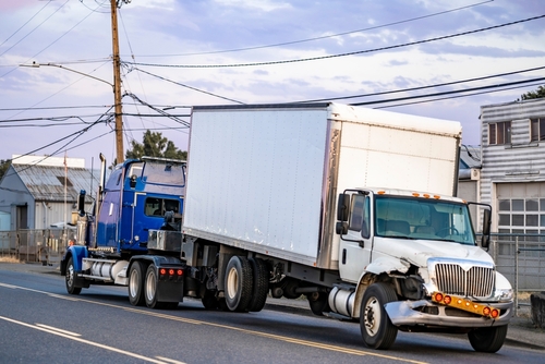 A damaged truck being towed with visible front-end damage, indicating it was involved in a serious accident on a road lined with utility poles and buildings.