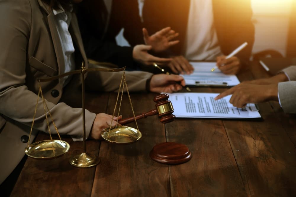 A judge's gavel placed on a desk with a team of lawyers engaged in a meeting in the background, symbolizing collaboration and legal services within a law firm.