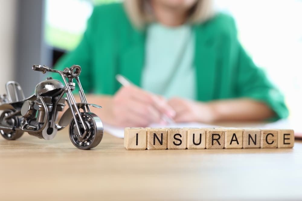 Motorcycle travel and medical insurance concept: a motorcycle model and the word "insurance" displayed on a table, with a blurred manager reviewing documents in the background.