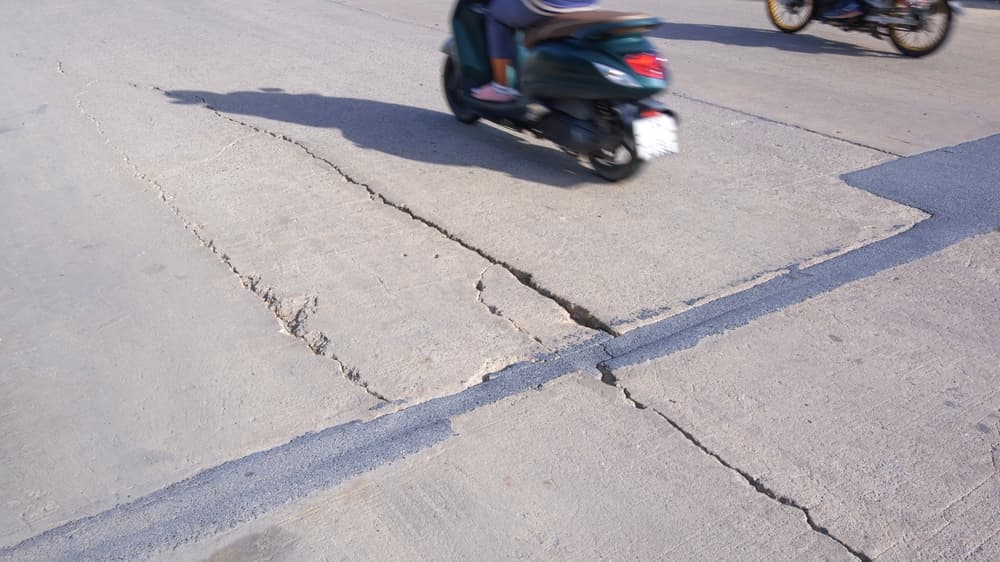 Two motorcycles in motion blur, speeding along an old, damaged concrete road. The road surface is visibly worn, with long, jagged crack lines and areas of collapse that add a rugged texture.