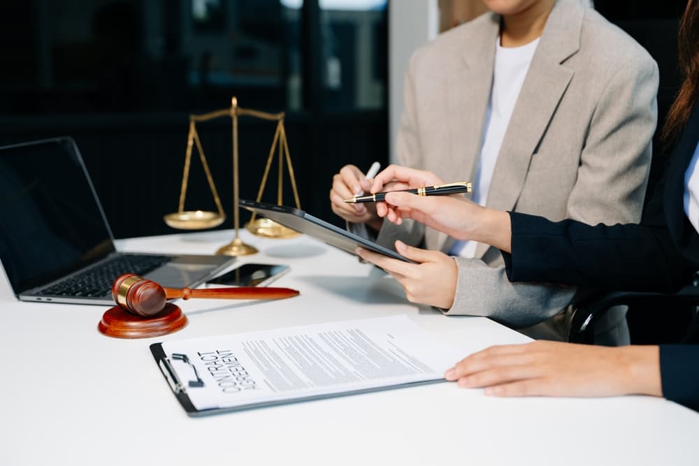 Lawyers and business professionals reviewing contract documents in an office, with a brass scale of justice prominently displayed on the desk. 