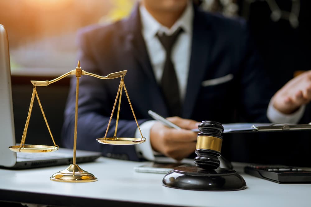 A judge's gavel placed on a desk alongside legal documents, with a lawyer reviewing paperwork in the background. This scene symbolizes legal advice, justice, and the practice of law.


