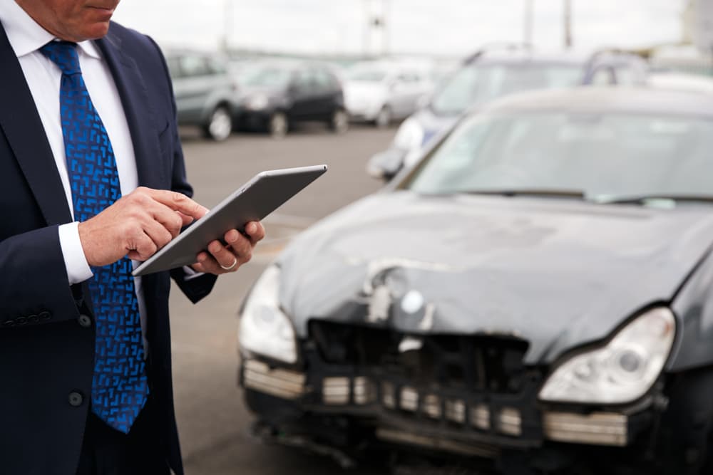 Male insurance adjuster with a digital tablet assessing car damage after a motor accident.