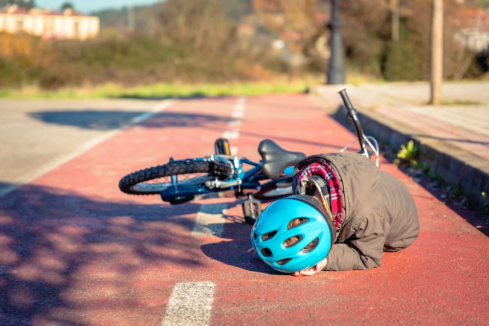 A boy sitting on the ground in the street, holding his helmeted head after falling off his bicycle.