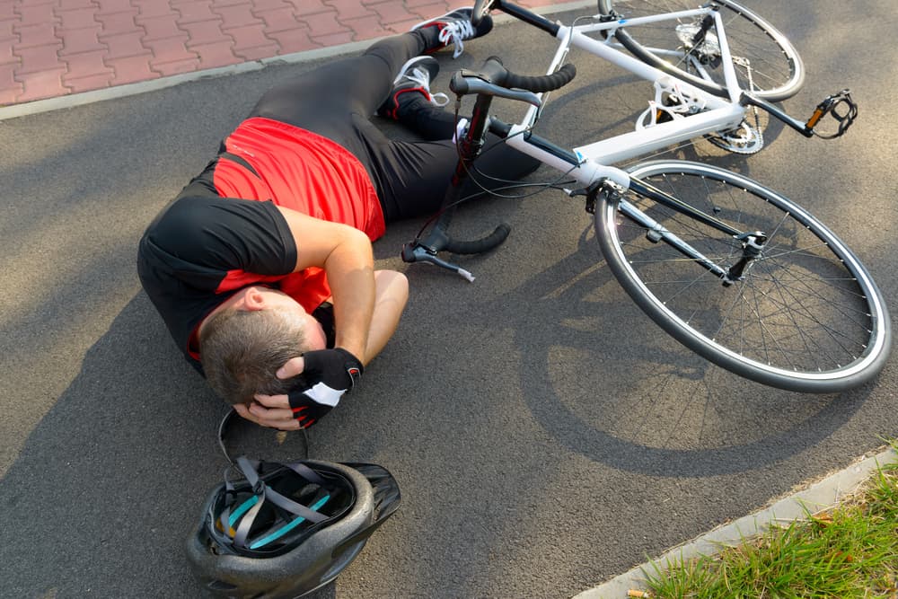 A cyclist lies on the road, clutching their head, showing signs of a severe traumatic brain injury following a bicycle accident.