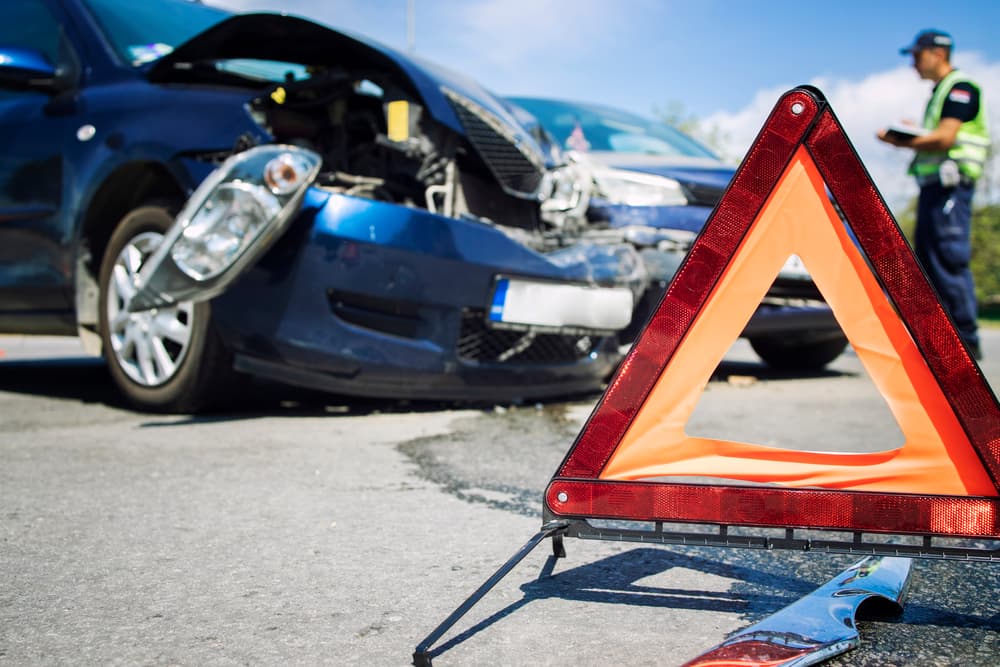 A car accident at a busy intersection. Vehicles are damaged and a warning triangle is set up to alert traffic. In the background, a police officer is seen documenting the accident report.