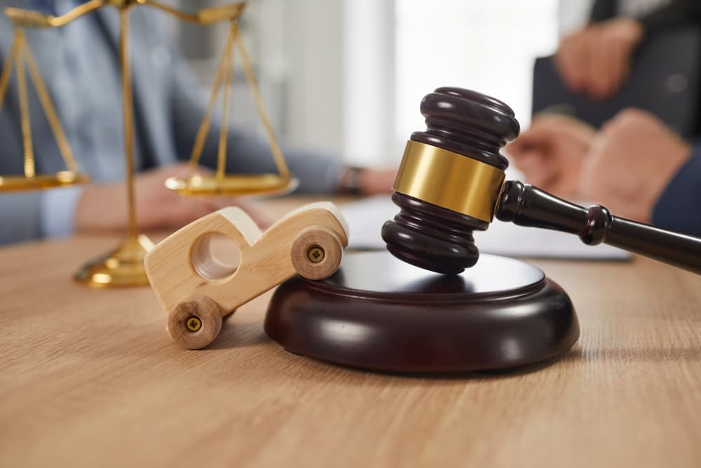 A small wooden toy car, sound block, gavel, and scales of justice are placed on an office table, symbolizing legal services and accident law.