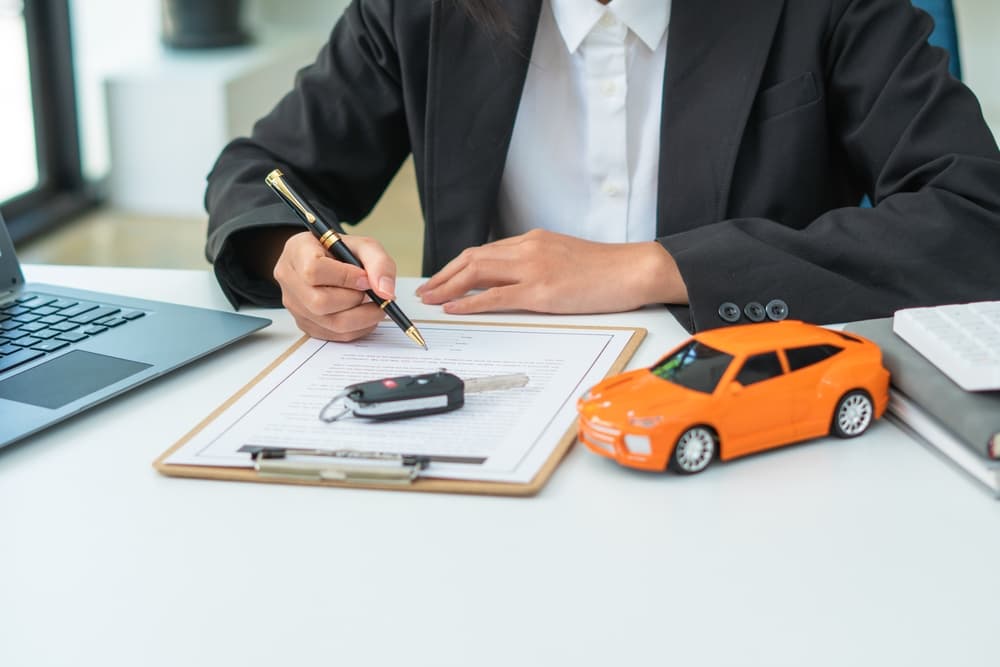 A man signs a car insurance document or lease agreement, his signature flowing across the contract. Car keys rest on the table beside him, symbolizing the purchase or sale of a new or used vehicle.