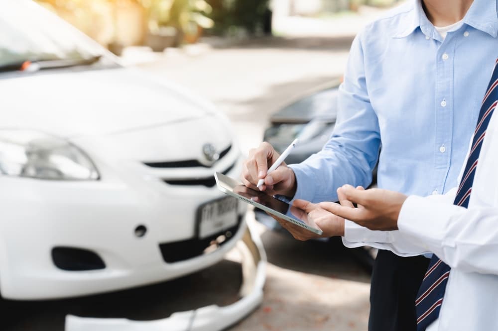 An insurance agent inspects a damaged car while assisting with the claim process after a crash. The customer submits a digital signature on a tablet to complete the claim report, highlighting the car insurance and accident settlement process.







