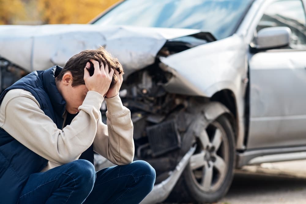 A distressed man stands near his wrecked car, His face shows pain and confusion as he clutches his head, feeling intense pain from a severe head injury after the accident.