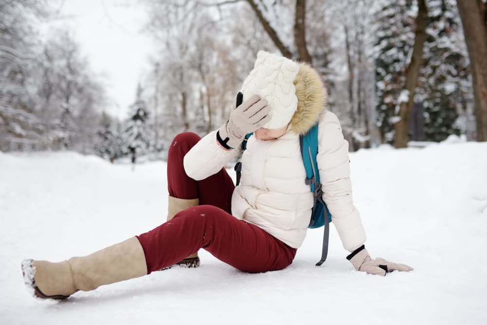 A woman slipping on an icy path in a snowy winter park, falling and lying on the ground, highlighting the danger of seasonal injuries.