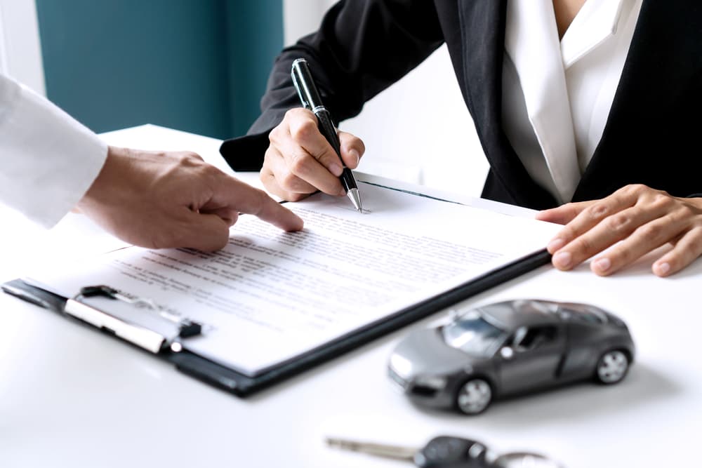 Close-up of an Asian woman signing a car insurance document, lease agreement, or sales contract. Car keys rest on the table, symbolizing the purchase or sale of a new or used vehicle.