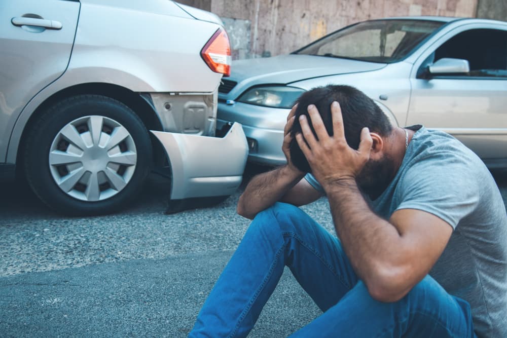 A man standing near a car accident, clutching his head in pain.