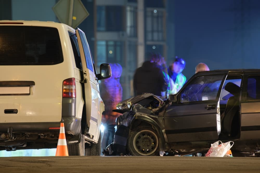 A heavy car crash on a city street at night, with silhouettes of people standing nearby. The scene reflects road safety and insurance concerns.