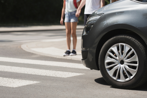 A car approaches a crosswalk where pedestrians are standing on the curb, indicating a potential interaction between drivers and pedestrians.