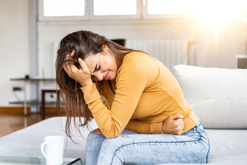 A young woman sits on a couch, holding her head and stomach in visible discomfort, portraying pain and emotional distress.