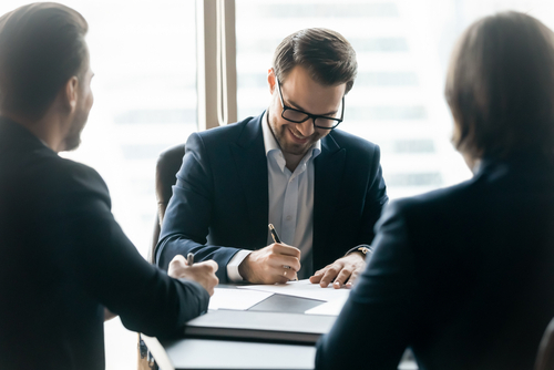 A lawyer in a suit smiles while signing documents during a meeting with two other professionals, representing legal consultation or agreement.