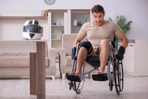 A man in a wheelchair wearing a knee brace smiles energetically, with a motorcycle helmet placed on a nearby table in a home setting.