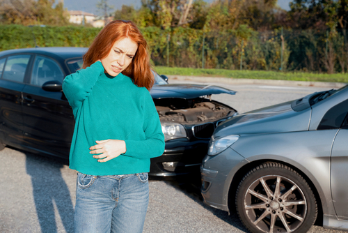 A red-haired woman holds her neck in pain while standing near two cars involved in a rear-end collision, indicating potential injury.