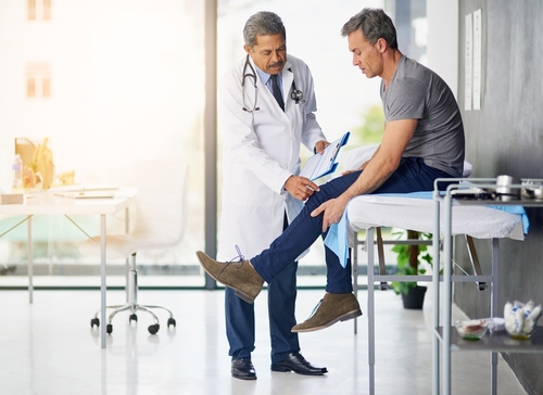 A doctor examines a man’s injured leg while he sits on an examination table in a bright medical office.