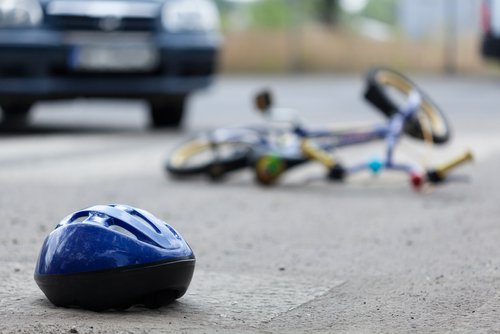 A blue bicycle helmet lies on the ground in focus, with a bicycle and a car blurred in the background, depicting a potential bicycle accident.