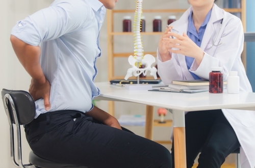 A patient holding their lower back consults with a doctor at a desk, with a model of the spine in the background, indicating a spinal cord injury discussion.