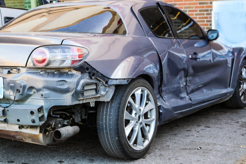 A heavily damaged silver car, still drivable, is parked at a store. The right rear side shows visible impact from a collision.