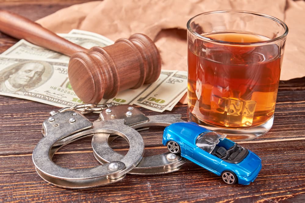 A judge's gavel, a full glass of whiskey, handcuffs, a miniature car, and dollar bills on a wooden background—symbolizing the legal and financial consequences of drunk driving.