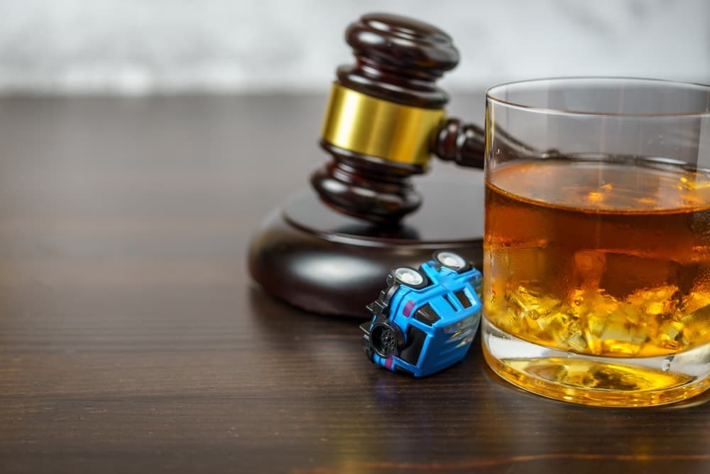 A judge's gavel on a courtroom table alongside a glass of whiskey and a small toy car, symbolizing the dangers of drinking and driving.